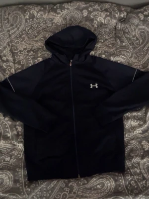 Mörkblå hoodie från Under Armour - Snygg mörkblå hoodie från Under Armour med dragkedja och huva. Framtill finns en vit logga på bröstet och på ryggen står det 'Under Armour' i vitt. Perfekt för träning eller chill. Materialet känns sportigt och bekvämt.