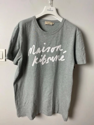 Maison Kitsuné t-shirt - Snygg grå t-shirt från Maison Kitsuné med vit logotyptext framtill. Klassisk rund halsringning och korta ärmar. Tillverkad i mjuk bomull för en skön och avslappnad look. Perfekt till jeans eller shorts. L fits M
