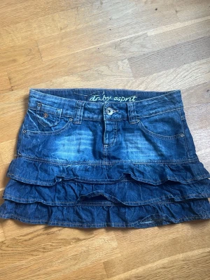 Blå jeanskjol med volanger  - Säljer en blå jeanskjol från Esprit med volanger och klassiska fickor både fram och bak. Kjolen har bälteshällor och knappstängning framtill. Perfekt för dig som gillar en avslappnad men ändå söt stil.