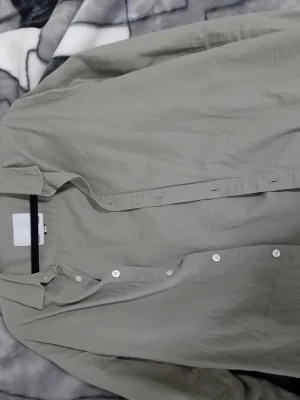 Beige skjorta från 157 - Klassisk beige skjorta från 157 i storlek S. Skjortan har en rak passform, långa ärmar och vita knappar framtill. Tillverkad i mjukt bomullstyg som känns skönt mot huden. Perfekt att styla lager på lager eller bära som den är.