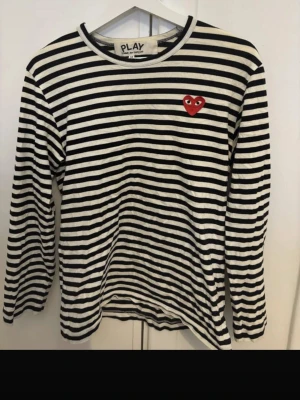 Randig långärmad topp från Comme des Garçons - Svartvit randig långärmad topp från Comme des Garçons PLAY med det ikoniska röda hjärtat broderat på bröstet. Mjuk bomull och klassisk passform, perfekt för en stilren look. Sitter som S typ!