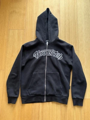 Svart zip hoodie från Hoodrich - Säljer en svart hoodie med dragkedja från Hoodrich. Hoodien har stor huva, två fickor framtill och vit broderad logga över bröstet. Tillverkad i mjukt bomullsmaterial och har ribbade muddar vid ärmar och nederkant. Perfekt för en chill och street-inspirerad look.