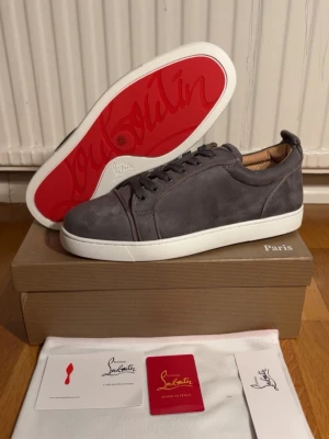 Gråa Christian Louboutin sneakers - Snygga gråa sneakers från Christian Louboutin i mocka med klassisk röd sula och vita kanter. Lågt skaft och diskret design med ton-i-ton snörning. Tillverkade i Italien och levereras med originalkartong och dustbag.