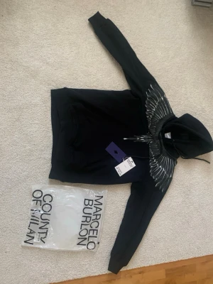 Svart hoodie Marcelo Burlon vingar - Säljer en svart hoodie från Marcelo Burlon County of Milan med ikoniskt vingtryck i vitt och grått över axlar och bröst. Hoodien har huva med dragsko och stor magficka framtill. Materialet är mjuk bomull som känns riktigt skön mot huden.