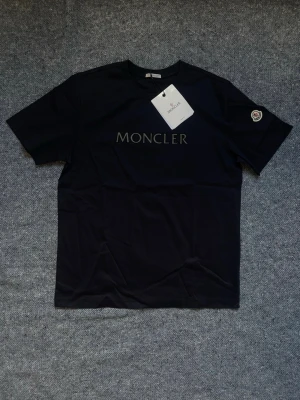 Navy Moncler t-shirt med logga - Snygg svart t-shirt från Moncler med klassisk logga tryckt på bröstet och Moncler-märke på ärmen. T-shirten har rund hals och korta ärmar, perfekt för en clean och stilren look. Tillverkad i mjuk bomull för maximal komfort.Finns även i svart och beige. Storlek L