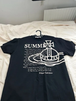 Svart t-shirt Vivienne Westwood - Svart t-shirt från Vivienne Westwood med stort vitt tryck framtill som inkluderar text, grafisk hjälm och William Shakespeare-citat. Klassisk rund hals och korta ärmar. Perfekt statement-plagg för dig som gillar unika designer.