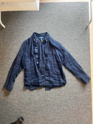 Marinblå linneskjorta från Polo Ralph Lauren - Säljer en marinblå skjorta från Polo Ralph Lauren i svalt linne. Skjortan har klassisk krage, vita knappar och den ikoniska röda logotypen broderad på bröstet. Perfekt för dig som gillar en avslappnad men ändå stilren look. Det är barn 14-16 år men det är som xs