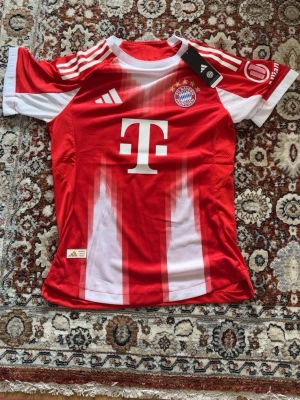 Bayern München matchtröja Adidas - Röd och vit Bayern München matchtröja från Adidas med klubbmärke, sponsorlogga och nummer 17 Olise på ryggen. Tröjan har korta ärmar, rund hals och är tillverkad i ett lätt, ventilerande funktionsmaterial. Perfekt för fotbollsträning eller match.