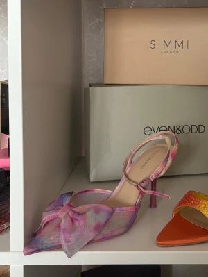 Pastellfärgad pumps med rosett - Trendig pumps från Even&Odd i pastellfärgat tyg med stora rosa, lila och blå toner. Skon har en spetsig tå, öppen häl och en stor dekorativ rosett framtill. Justerbar rem runt ankeln 