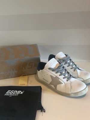 Golden goose - Säljer ett par vita Golden Goose sneakers med grå stjärna på sidan, grå skosnören och mörkblå detalj vid hälen. Skorna har en klassisk låg siluett och är tillverkade i läder med slitna detaljer för en cool, avslappnad look. Perfekta för dig som gillar streetstyle.