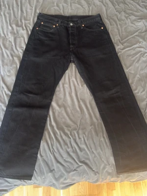 Svarta raka levi buksar - Säljer ett par svarta jeans med klassisk rak passform och fem fickor. Jeansen har knappgylf och nitar i kopparfärg vid fickorna. Tillverkade i kraftig denim, perfekta för en stilren och enkel look.