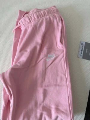 Rosa mjukisbyxor från Nike - Snygga ljusrosa mjukisbyxor från Nike med broderad vit logga på benet. Byxorna har dragsko i midjan och är gjorda i mjukt bomullsmaterial för maximal komfort. Perfekta för chill dagar eller när du vill ha en avslappnad look.
