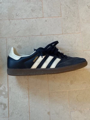 Adidas Samba sneakers marinblå - Klassiska Adidas Samba sneakers i marinblått med vita detaljer och bruna gummisulor. Skorna har de ikoniska tre ränderna på sidan och guldfärgad Samba-text. Ovandelen är i läder och mocka, med lågt skaft och snörning. Perfekta för en sportig och stilren look. Bra skick svåra o få tag på i denhär storleken 