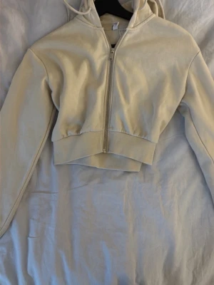 Beige cropped hoodie med dragkedja - Snygg beige cropped hoodie med dragkedja framtill och huva med dragsko. Tröjan har långa ärmar och bred ribbad mudd i midjan för en modern look. Perfekt att slänga på sig över ett linne eller t-shirt för en avslappnad stil.