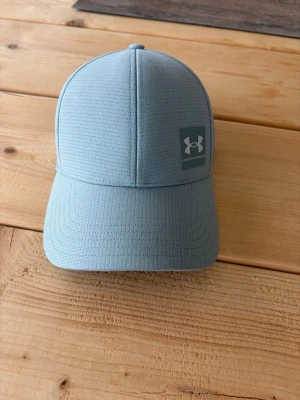 Ljusblå keps från Under Armour S/M - Snygg ljusblå keps från Under Armour med böjd skärm och diskret rutmönster i materialet. Kepsen har ett stilrent logotypmärke framtill och är tillverkad i ett lätt, ventilerande syntetmaterial som passar perfekt till sport och fritid.
