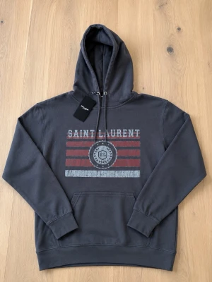 Saint Laurent hoodie - Snygg mörkgrå hoodie från Saint Laurent med tryck framtill i vitt och vinrött. Klassisk känguruficka, justerbar huva med snören och ribbade muddar. Perfekt för dig som vill ha en stilren och trendig look. Tillverkad i mjuk bomull.