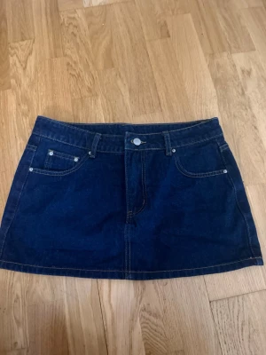 Mörkblå jeanskjol i denim - Snygg mörkblå jeanskjol i klassisk denim med fem fickor, bälteshällor och knappstängning fram. Kjolen har en rak och kort modell med tydliga sömmar och silverfärgade nitar. Perfekt för dig som gillar en enkel men trendig look.