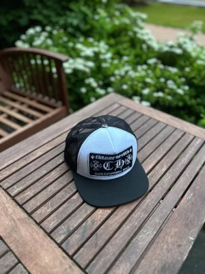Chrome Hearts svart och vit keps - Säljer en snygg svart och vit keps från Chrome Hearts med mesh på sidorna och bak. Framsidan har ett stort broderat patch med logotyp och text. Skärmen är svart och kepsen har justerbar snapback. Perfekt för dig som vill sticka ut med en cool accessoar.