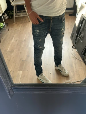 Blå slitna skinny jeans - Säljer ett par blå skinny jeans med slitningar och hål på framsidan. Jeansen har klassisk femficksdesign och detaljerade slitningar även på bakfickorna. Sitter tight och har en modern streetstil som passar perfekt till sneakers.