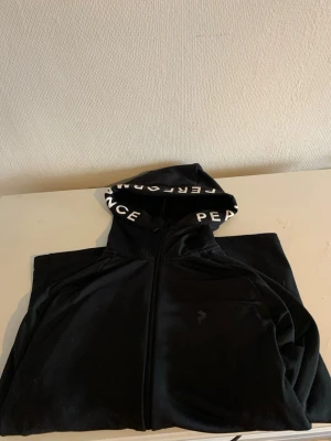 Peak performance zip hoodie  - Snygg svart hoodie från Peak Performance med dragkedja och huva. På huvan står det 'PERFORMANCE' och 'PEAK' i vitt. Perfekt för en avslappnad och sportig stil. Materialet känns mjukt och bekvämt.