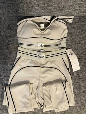 Beige träningsset från Alo Yoga - Snyggt beige träningsset från Alo Yoga med svarta kontrastsömmar. Setet består av en sport-bh och matchande tights i stretchigt funktionsmaterial. Perfekt för yoga, gym eller annan träning. Elastisk midja med Alo-logga och modern design.