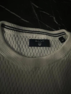 Vit stickad tröja från Gant - Klassisk vit stickad tröja från Gant med rund halsringning och diskret logga broderad på bröstet. Tröjan har lång ärm och ribbade muddar vid ärmslut och nederkant. Tillverkad i mjukt material som ger en clean och stilren look.