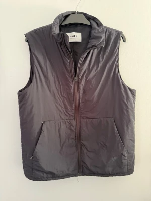 Svart dunväst från NN07 - Stilren svart dunväst från NN07 med dragkedja framtill och två stora fickor. Västen har hög krage och justerbar dragsko i nederkant. Perfekt att slänga över en hoodie eller tröja för extra värme. Materialet är glansigt och lätt, vilket gör den enkel att bära. Den har defekter som man ser på bilderna!