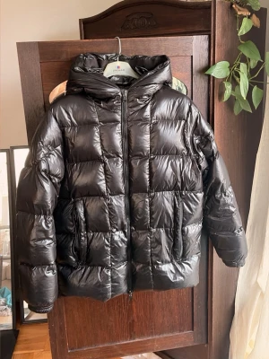 Moncler jacka  - Snygg svart dunjacka från moncler med glansig finish och stor huva. Jackan har dragkedja framtill och två sidofickor. Perfekt för kalla vinterdagar och riktigt varm tack vare dunfyllningen.