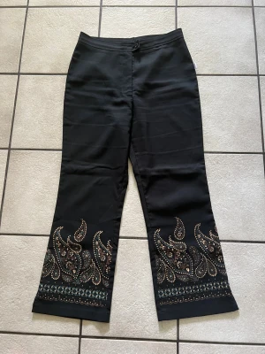 Svarta 3/4 bootcut byxor med paisleymönster storlek L - Unika svarta bootcut byxor med färgglatt paisleymönster och detaljer längst ner på benen. Perfekt för dig som vill sticka ut med coola mönster och snygg passform. Byxorna har knapp och dragkedja framtill och är tillverkade i ett mjukt material. Midja 70 cm