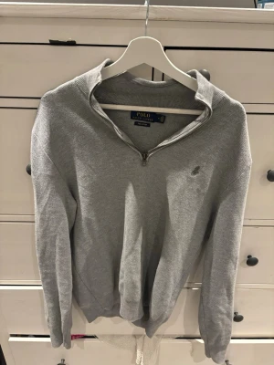 Grå halvzip från Polo Ralph Lauren - Snygg grå stickad tröja från Polo Ralph Lauren i mjuk pima cotton. Tröjan har halv dragkedja, ribbade muddar och diskret logga på bröstet. Perfekt till jeans eller chinos för en clean look.