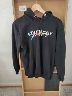 Svart Givenchy hoodie med färgglad logga - Säljer en svart hoodie från Givenchy med en färgglad broderad logga på bröstet där det står 'Givenchy Paris'. Hoodien har huva med vita öljetter och ribbade muddar vid ärmslut och nederkant. Perfekt för dig som gillar streetwear och vill sticka ut.