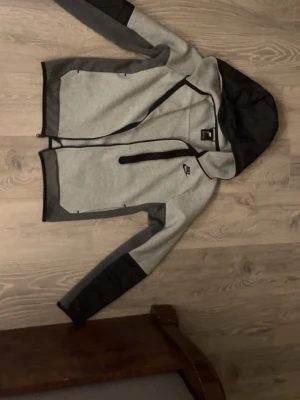 Grå Nike hoodiejacka med svarta detaljer - Snygg grå hoodiejacka från Nike med svarta detaljer på ärmar, huva och dragkedja. Jackan har en klassisk Nike-logga på bröstet och praktisk huva. Tillverkad i mjukt material som passar perfekt till streetwear och vardagsstil.