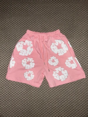 Rosa shorts denim tear - Säljer ett par rosa shorts med vita, upphöjda blommönster och elastisk midja med snörning. Shortsen har en avslappnad passform och är perfekta för varma dagar. Materialet känns mjukt och bekvämt, troligen bomull eller polyester.
