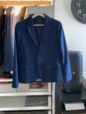 Mörkblå blazer från Tommy Hilfiger - Snygg mörkblå blazer från Tommy Hilfiger med klassisk passform och två knappar framtill. Kavajen har två fickor med lock, bröstficka och är helfodrad. Perfekt för en stilren look och passar till både skjorta och t-shirt. Nypris: 1800kr