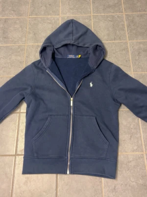 Blå Ralphlauren zip hoodie - Snygg blå hoodie från Polo Ralph Lauren med dragkedja framtill och klassisk vit logga broderad på bröstet. Hoodien har två stora fickor fram och mjukt fleecefoder på insidan. Perfekt för en avslappnad och stilren look.