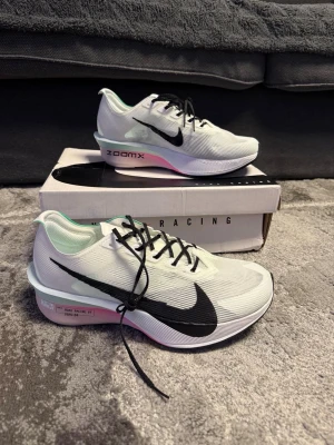 Nike ZoomX Vaporfly NEXT% sneakers - Nike ZoomX Vaporfly NEXT% sneakers i vitt med svart swoosh, svarta skosnören och detaljer i grönt och rosa. Ovandelen är i mesh för bra ventilation och sulan har ZoomX-dämpning för extra studs. Perfekta för dig som vill ha lätta och snabba sneakers.