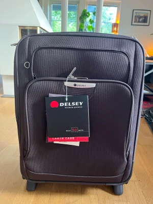 Svart kabinväska från Delsey - Snygg svart kabinväska från Delsey med två hjul, TSA-lås och praktiska ytterfack. Väskan har dragkedja och är tillverkad i slitstarkt syntetmaterial. Perfekt storlek och enkel att rulla genom flygplatsen.