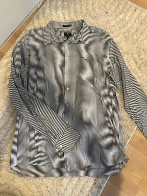 Randig skjorta från GANT, blå/vit - Säljer en klassisk blå- och vitrandig skjorta från GANT i modellen Tech Prep Oxford Slim. Skjortan har lång ärm, knappar framtill och en liten broderad logga på bröstet. Tillverkad i bomull för en skön och luftig känsla.