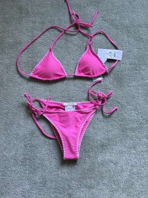 Rosa triangelbikini med vita detaljer - Snygg rosa bikini med triangelöverdel och matchande trosa. HELT NY!!💕 Fick när jag fyllde men vi hann inte lämna  tillbaka den inom 14 dar och behöver en större storlek❤️