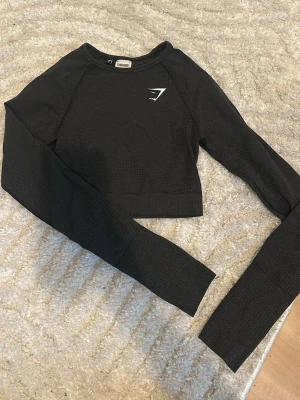 Svart långärmad crop top från Gymshark - Snygg svart långärmad crop top från Gymshark med diskret mönster i tyget och vit logga på bröstet. Tight passform och stretchigt material som passar perfekt till träning eller gym. Rund halsringning och sömlös design för extra komfort.