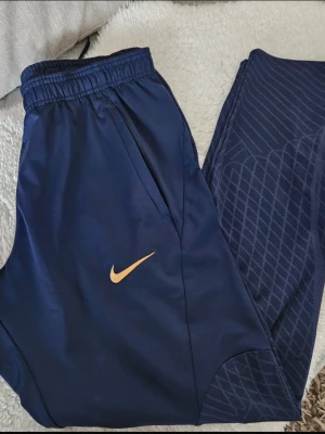 Marinblå Nike PSG träningsbyxor - Snygga mörkblå träningsbyxor från Nike med Paris Saint-Germain-logga och guldfärgad . Byxorna har elastisk midja med dragsko, fickor och diskret mönster på benen. Perfekta för träning eller chill. Tillverkade i ett mjukt och stretchigt material.