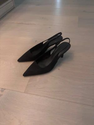 Svarta slingback pumps med låg klack - Stilrena svarta pumps med spetsig tå och slingback-rem. Skorna har låg klack och är tillverkade i mockaimitation med innersula i syntet. Perfekta för en chic och elegant look.