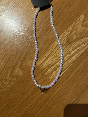 Pärlhalsband med hängsmycke - Snyggt halsband med vita pärlor och ett litet hängsmycke i form av ett djur. Halsbandet har en klassisk och stilren look med rundade pärlor som ger en elegant känsla. Perfekt accessoar för att lyfta din outfit.