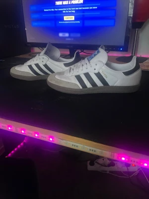 Adidas Samba vita och svarta sneakers - Klassiska Adidas Samba sneakers i vitt läder med svarta ränder och grå mockadetaljer vid tån. Gummisula och låg profil. Perfekta för dig som gillar retrostil och streetwear. Ikonisk Adidas-logga på plösen och sidan.