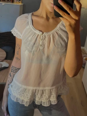 Topp från abercrombie and fitch - Söt och luftig vit blus med korta ärmar, spetsdetaljer vid ärmslut och nederkant samt volang längst ner. Blusen har rund halsringning med knappar framtill och broderade blommor vid fållen. Från abercrombie and fitch storlek s