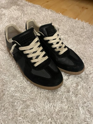 Maison Margiela svarta sneakers - Köpte från minito Maison Margiela sneakers i svart läder och mocka med beigea skosnören och klassisk gummisula. Ikonisk tab på plösen med siffror och logga. Snygg retrostil som passar till många outfits.