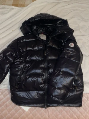 Moncler maya  - Snygg svart dunjacka från Moncler med glansig finish, huva och två dragkedjeförsedda fickor. Jackan har en klassisk Moncler-logga på ärmen och quiltad design som ger en puffig look. Perfekt för kalla vinterdagar.