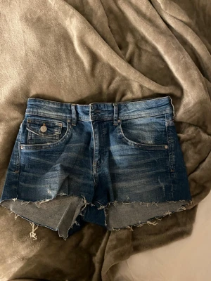 Blå jeansshorts - Säljer ett par blå jeansshorts med fransig, rå kant och klassisk femficksdesign. Shortsen har normal passform och är tillverkade i slitstarkt denimtyg. Perfekta för varma dagar och en avslappnad stil.