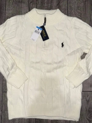 Vit kabelstickad tröja Polo Ralph Lauren - Kabelstickad vit tröja från Polo Ralph Lauren med halv dragkedja och ribbade muddar. Klassisk svart broderad logga på bröstet. Tröjan har hög krage och är gjord i mjukt material, perfekt för kyliga dagar.