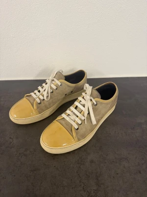 Beige och gula sneakers med lackdetalj från Lanvin - Sneakers i beige mocka med vita snören och gulglänsande lack på tån. Klassisk låg modell med vit sula och svarta detaljer på insidan. Snygg kontrast mellan material och färger som ger en unik look. Skorna är i storlek 43